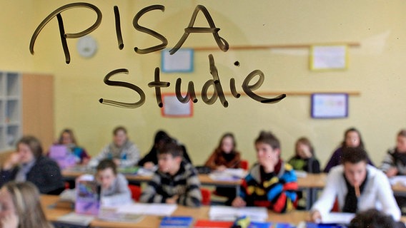 Die Wörter "PISA Studie" stehen auf einer Glasscheibe in einer Schulklasse, während Schüler den Unterricht verfolgen. © dpa Foto: Julian Stratenschulte