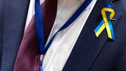 Ein Kabinettsmitglied trägt eine Schleife in den Farben der ukrainischen Nationalflagge während eines Gesprächs der EU-Außenminister am runden Tisch im Gebäude des Europäischen Rates. © AP/dpa Foto: Virginia Mayo
