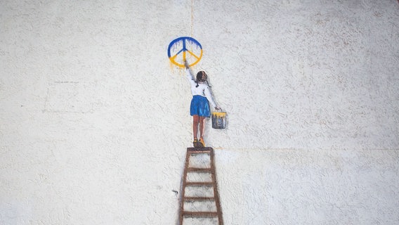 Ein Kunstwerk des italinischen Street-Art Künstlers Salvatore Benintende Tvboy in Butcha zeigt ein Mädchen auf einer Leiter, das ein blau-gelbes Peace-Zeichen an die Wand malt. © picture alliance Foto: Oleksii Chumachenko