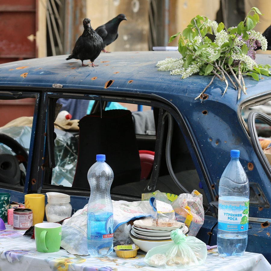 Tauben sitzen auf einem zerstörten Auto und suchen nach Essen, davor steht ein Tisch mit Geschirr und Essensresten. © Anadolu Agency Foto: Leon Klein