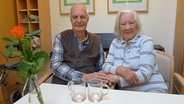 Ein älteres Paar sitzt in einem Pflegeheim Händchen haltend nebeneinander. © NDR Foto: Bettina Less