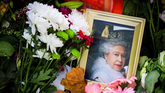 London: Ein Foto von Königin Elizabeth II. und andere Trauerbekundungen liegen zwischen Blumen auf einer Wiese im Green Park neben dem Buckingham-Palast. © dpa Foto: Christian Charisius