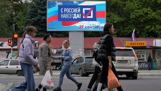 Menschen gehen an einer elektronischen Anzeige mit der Aufschrift "Für immer mit Russland" in der von Russland unterstützten und von Separatisten kontrollierten Volksrepublik Luhansk vorbei. © Uncredited/AP/dpa 