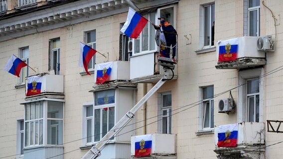 Arbeiter hängen russische Fahnen an einem Wohnhaus in der von Russland unterstützten und von Separatisten kontrollierten Volksrepublik Luhansk auf. © Uncredited/AP/dpa 
