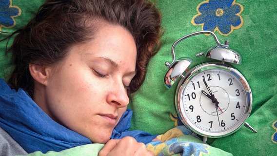 Eine Frau liegt im Bett und schläft, neben ihr liegt ein Wecker auf dem Kopfkissen. © picture alliance 