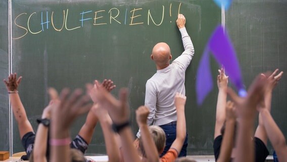 Ein Lehrer schreibt mit bunder Kreide an eine Tafel das Wort "Schulferien!!". Im Vordergrund sind die jubelnden Arme von Schülern zu sehen. © picture alliance / Daniel Karmann/dpa Foto: Daniel Karmann