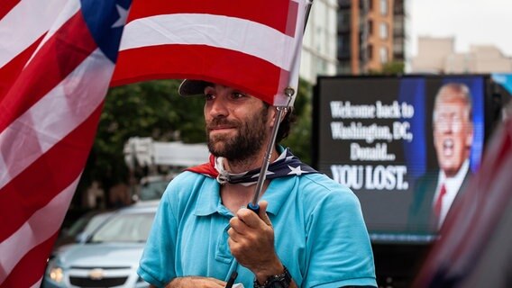 Ein Mann schwenkt eine US-Flagge, im Hintergrund ein Plakat-LKW mit Donald Trump. © picture alliance Foto: Allison Bailey