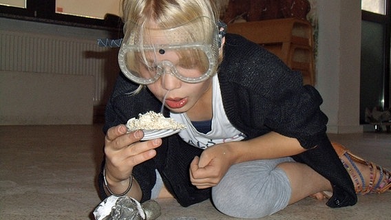 Ein Mädchen mit Schutzbrille pustet auf eine kleine Flamme in einer mit Sägespänen gefüllten Muschelschale. Ein Mädchen mit Schutzbrille pustet auf eine kleine Flamme in einer mit Sägespänen gefüllten Muschelschale. Foto: Aline König