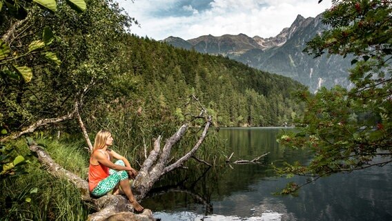 Eine Frau sitzt in lockerer Kleidung auf einem Baumstamm und schaut auf den Pigurber See im Naturpark Öztal © Oesterreich Werbung / The Creating Click.com 