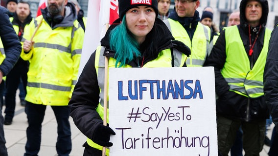 Eine Mitarbeiterin hält ein Schild mit der Aufschrift Lufthansa #SayYesTo Tariferhöhung bei einer Kundgebung der Unternehmenssparte Lufthansa Technik vor der Abflughalle im Terminal 1 des Flughafens BER. © Carsten Koall/dpa 