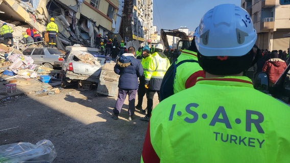 Mitglieder eines Rettungseinsatzteams stehen in den und vor den Trümmern eines bei einem Erdbeben in der Türkei zerstörten Gebäudes und suchen nach Überlebenden. © BRH-Rettungshundestaffel Hamburg und Harburg e.V. Foto: Grinnus