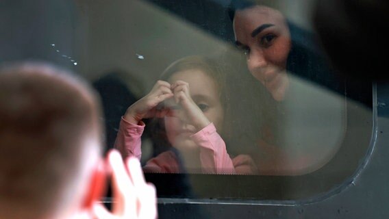 Eine Frau und ein Mädchen, das mit ihren Händen ein Herz formt, sitzen an einem Fensterplatz in einem Zug im Bahnhof von Lwiw. Auf dem Bahhsteig steht ein Mann und winkt ihnen. © dpa picture alliance/ASSOCIATED PRESS Foto: Daisuke Tomita