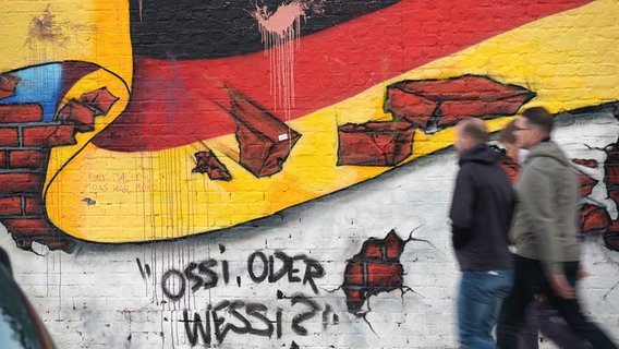 Berlin: Passanten gehen an einem Wandbild von Caspar Kirchner mit der deutschen Nationalflagge und dem Schriftzug "Ossi oder Wessi? vorbei. © dpa-Bildfunk Foto: Rainer Jensen/dpa