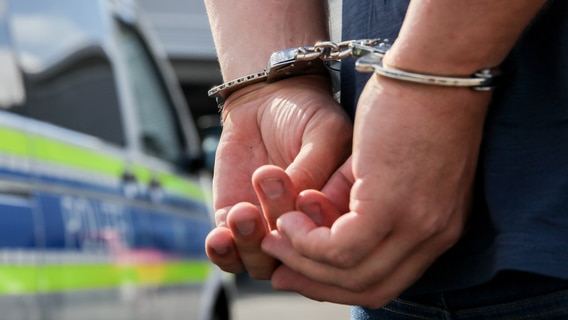 Ein Mann mit Handschellen steht vor einem Polizeiwagen. © picture alliance / Fotostand | Fotostand / K. Schmitt Foto: K. Schmitt