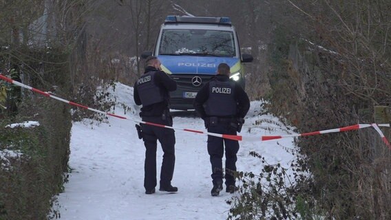 Polizisten laufen auf einem verschneiten Fußweg am Rand einer Kleingartenanlage auf ihr geparktes Auto zu, dessen Scheinwerfer noch angeschaltet sind. Im Vordergrund ist Absperrband zu sehen. © Felix Bänz Foto: Felix Bänz