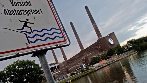 Im Vordergrund ist ein Schild mit der Aufschrift "Vorsicht Absturzgefahr" zu sehen, während im Hintergrund das VW-Werk zu sehen ist. © dpa - Bildfunk Foto: Julian Stratenschulte