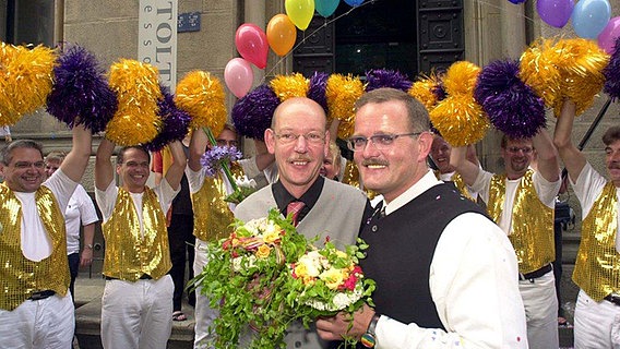 Heinz-Friedrich Harre (l.) und Reinhard Lüschow stehen mit Blumen vor einer Gruppe jubelnder männlicher Cheerleader. © NDR 