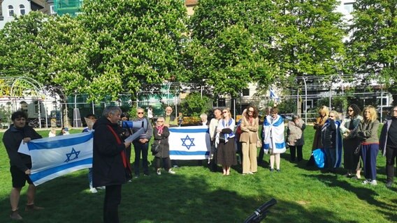 Eine Gruppe von rund 20 Frauen und Männern steht auf einem Rasen. Sie halten zwei Israel-Flaggen in den Händen, ein Mann hat ein Megaphon in der Hand und liest einen Text vor. © NDR Foto: Julia Willkomm