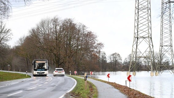 Ein Bus und ein Auto fahren an einem Hochwassergebiet vorbei, auf dem an Hochspannungsmasten das Wasser steht. © dpa Foto: Lars Penning