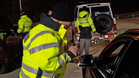 Ein Polizist kontrolliert auf einem Autobahnparkplatz die Papiere eines Autofahrers. © NDR 