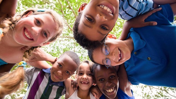 Eine Gruppe Kinder verschiedener Ethnien steht lächelnd im Kreis und schaut nach unten in die Kamera. © Imago Images Foto: Shotshop
