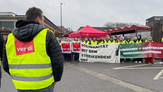 Ein Mann trägt eine gelbe Ver.di-Weste. Im Hintergrund mehrere Streikende mit Plakaten. © NDR Foto: Christian Nagel