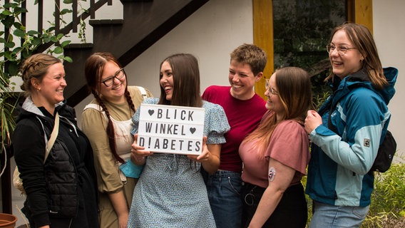 Mehrere Personen stehen in einer Gruppe und lachen sich an. Eine Person hält eine Tafel in der Hand, auf dieser steht: "Blickwinkel Diabetes" © NDR Foto: Julia Jänisch