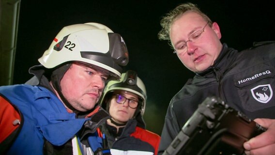 Drei Männer gucken auf einen Tablet-PC mit einer speziellen Feuerwehrapp bei Dunkelheit. © NDR 