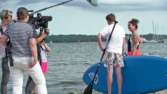 Ein Kamerateam und zwei Protagonist stehen mit SUP-Boards am Strand © NDR Foto: Lena Storm