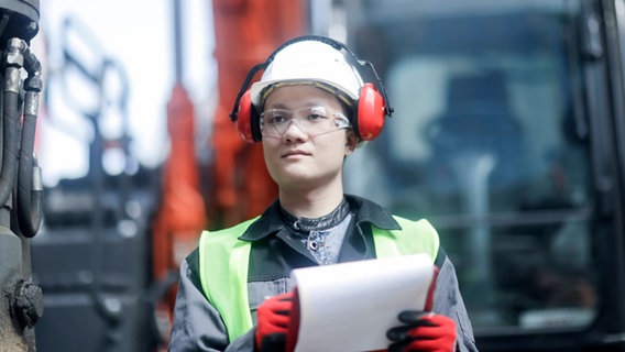 Eine Ingenieurin trägt einen Helm und Gehörschutz. Sie steht auf einer Baustelle und hält ein Klemmbrett. © IMAGO Foto: IMAGO / Cavan Images