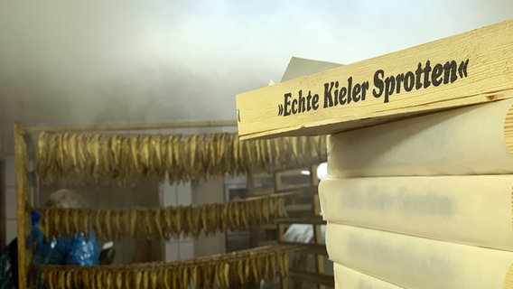 Eine Holzkiste mit der Aufschrift "Echte Kieler Sprotten" steht vorne im Bild, im Hintergrund hängen zahlreiche Fische auf einer Leine. © NDR Foto: Samir Chawki