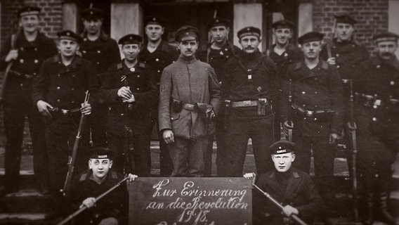 Eine Gruppe von Matrosen posieren für ein Foto hinter einem Schild: "Zur Erinnerung an die Revolution 1918". © NDR 