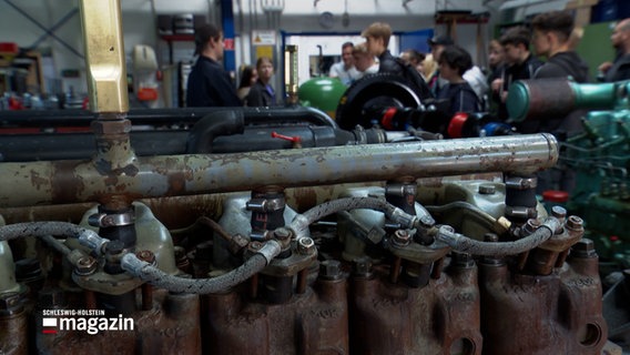 In Vordergrund ist ein Motor eines Schiffes, im Hintergrund bekommt eine Schulklasse eine Führung. © NDR Foto: NDR Screenshots
