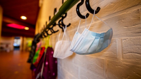 An einer Garderobe hängen zwei Masken im Hintergrund sind verschwommen noch Turnbeutel von Kindern an den Garderobenhacken zu erkennen. © xInderlied/Kirchner-Mediax 