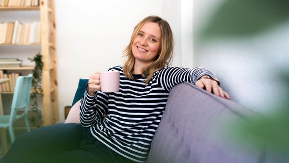 Eine lächelnde Frau sitzt mit einer Kaffeetasse auf einem Sofa. © IMAGO / Westend61 