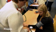 Eine Frau misst den Blutdruck bei einer Schülerin. © NDR Foto: NDR Screenshot