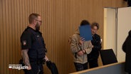 Ein Angeklagter wird in einem Gerichtssaal gebracht, er verdeckt sein Gesicht mit einer blauen Mappe. © NDR Foto: NDR Screenshot