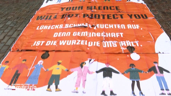 Ein Plakat auf dem Boden mit der Aufschrift: "Your Silence will not protect you - Lübecks Schulen leuchten auf, denn Gemeinschaft ist die Wurzel die uns hält. © NDR Foto: NDR Screenshot