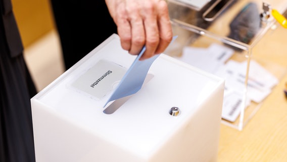 Eine Person wirft einen blauen Stimmzettel in eine weiße Box mit der Aufschrift Stimmzettel. © picture alliance / dpa Foto: Matthias Balk