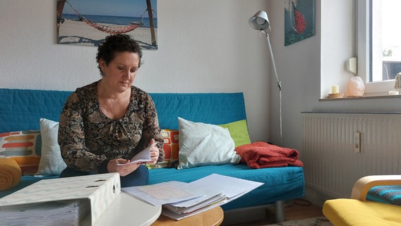 Eine Frau sitzt in ihrem Wohnzimmer und blickt auf einen Aktenordner mit Unterlagen. © NDR Foto: Hannah Böhme