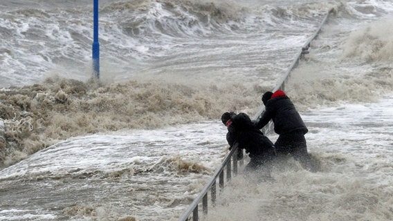 Zwei Männer halten sich an einem Geländer an der vom Sturm überspülten Uferpromenade in Dagebüll fest © picture alliance / dpa Foto: Carsten Rehder