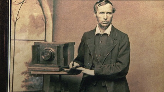 Ein Mann posiert neben einer historischen Kamera. © NDR 