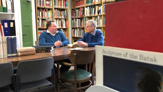 Zwei Männer sitzen sich in einer Bibliothek gegenüber. © NDR Foto: Philip Schroeder
