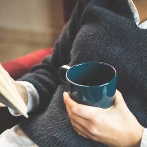 Eine junge Frau im blauen Pullover sitzt gemütlich im Sessel und hält einen Kaffeebecher und ein Buch in ihren Händen. © photocase.com Foto: Christine ten Winkel