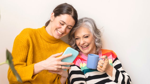 Eine jüngere und eine ältere Frau schauen gemeinsam auf ein Handy, dabei lachen sie. © IMAGO/Westend61 