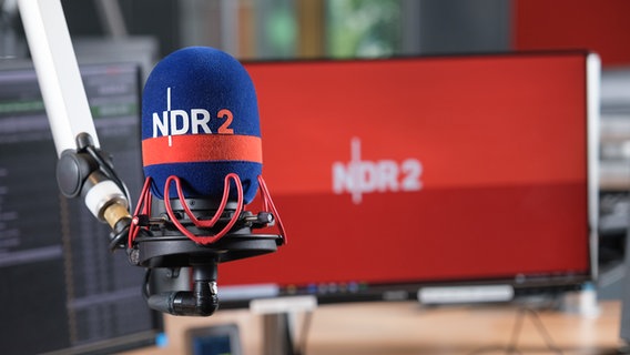 Blick auf ein Mikrofon mit NDR 2 Schützer im NDR 2 Studio mit einem Bildschirm im Hintergrund, auf dem auf rotem Grund ein weißes NDR 2 Logo zu sehen ist.  Foto: Niklas Kusche