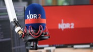 Blick auf ein Mikrofon mit NDR 2 Schützer im NDR 2 Studio mit einem Bildschirm im Hintergrund, auf dem auf rotem Grund ein weißes NDR 2 Logo zu sehen ist.  Foto: Niklas Kusche