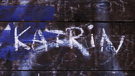 Mit Kreide auf die Holzwand einer alten Bushalte stelle gekritzelter Schriftzug "Katrin" - Motivbild zur NDR 2 Podcast-Reihe "Täter Unbekannt".  