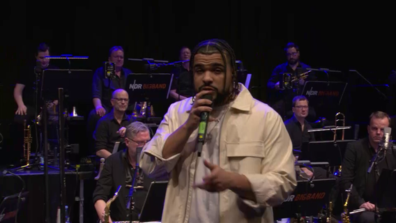 Screenshot: Leroy Menace am Mikrofon beim Konzert mit der NDR Bigband und TOYTOY auf Kampnagel. © NDR Bigband Foto: Screenshot