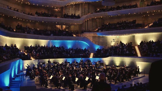 Screenshot: Das NDR Elbphilharmonie Orchester auf der Blau und Gelb beleuchteten Bühne des Großen Saals in der Elbphilharmonie Hamburg © NDR 